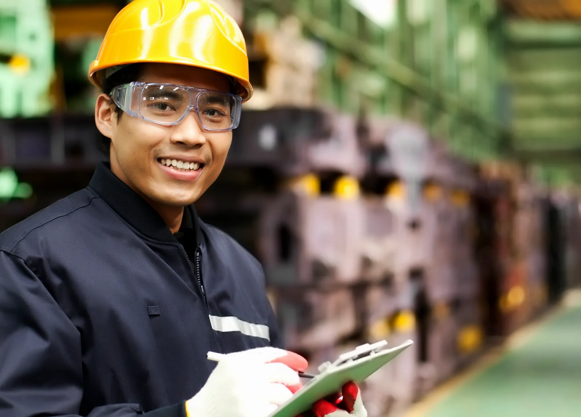 Smiling factory worker wearing safety gear and holding a clipboard.
