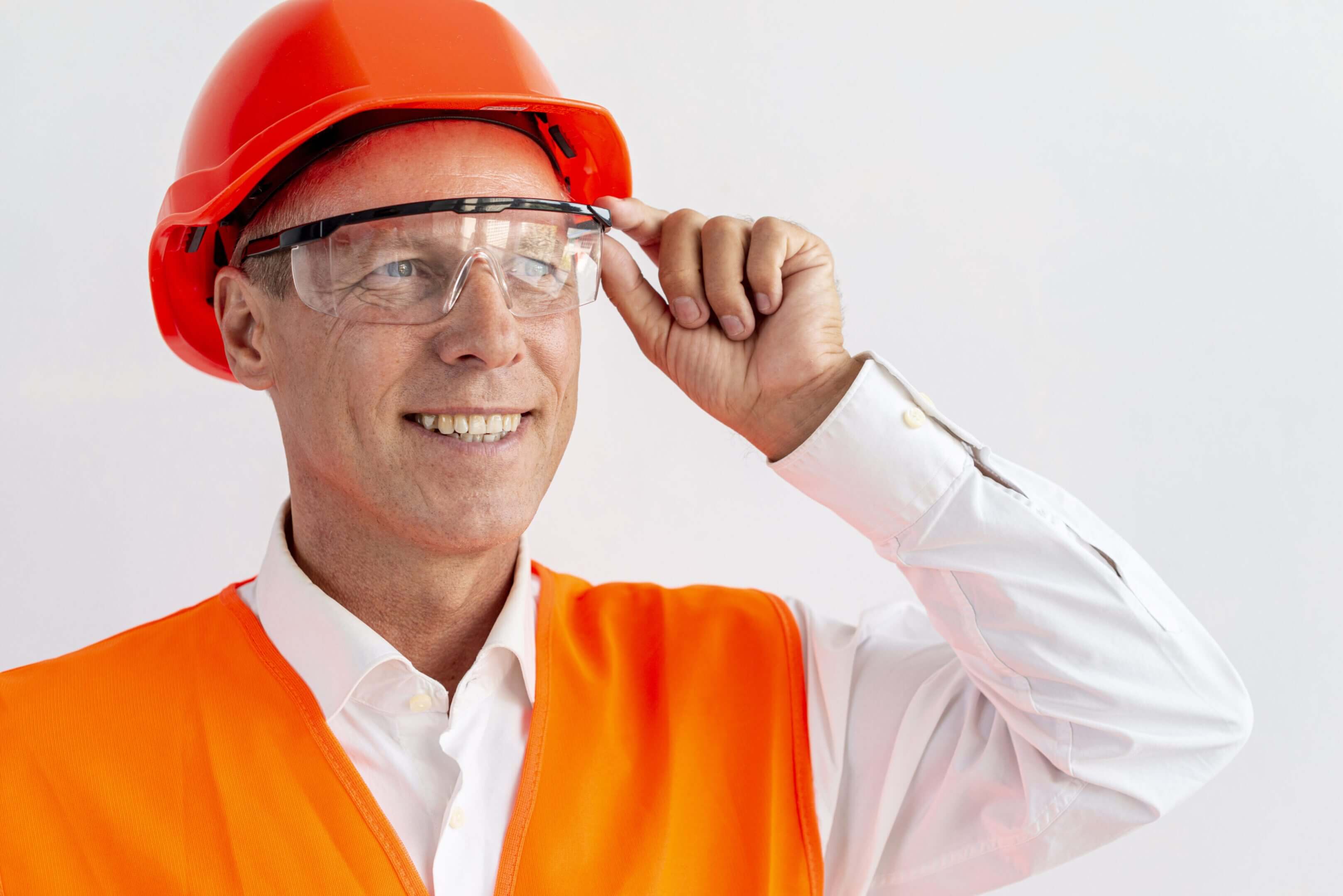 Smiling construction worker adjusting safety glasses and wearing an orange helmet and vest.